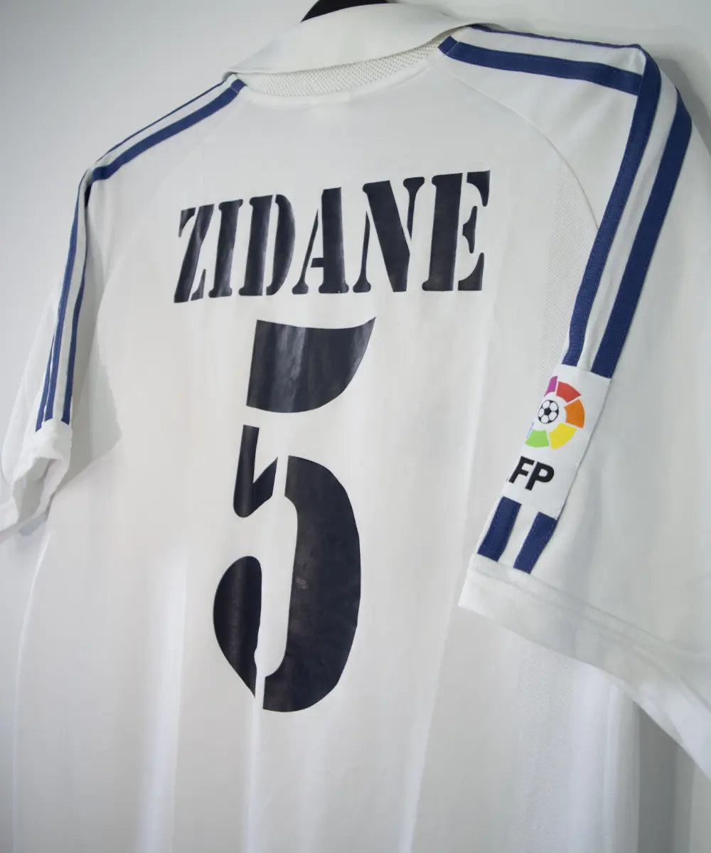 Maillot de foot vintage blanc domicile du real madrid de la saison 2001-2002. Il s'agit du maillot édité pour le centenaire. On peut retrouver l'équipementier adidas et le sponsor real madrid.com. Le maillot est floqué du numéro 5 Zidane. Le maillot comporte l'étiquette 695856 ASR001/09