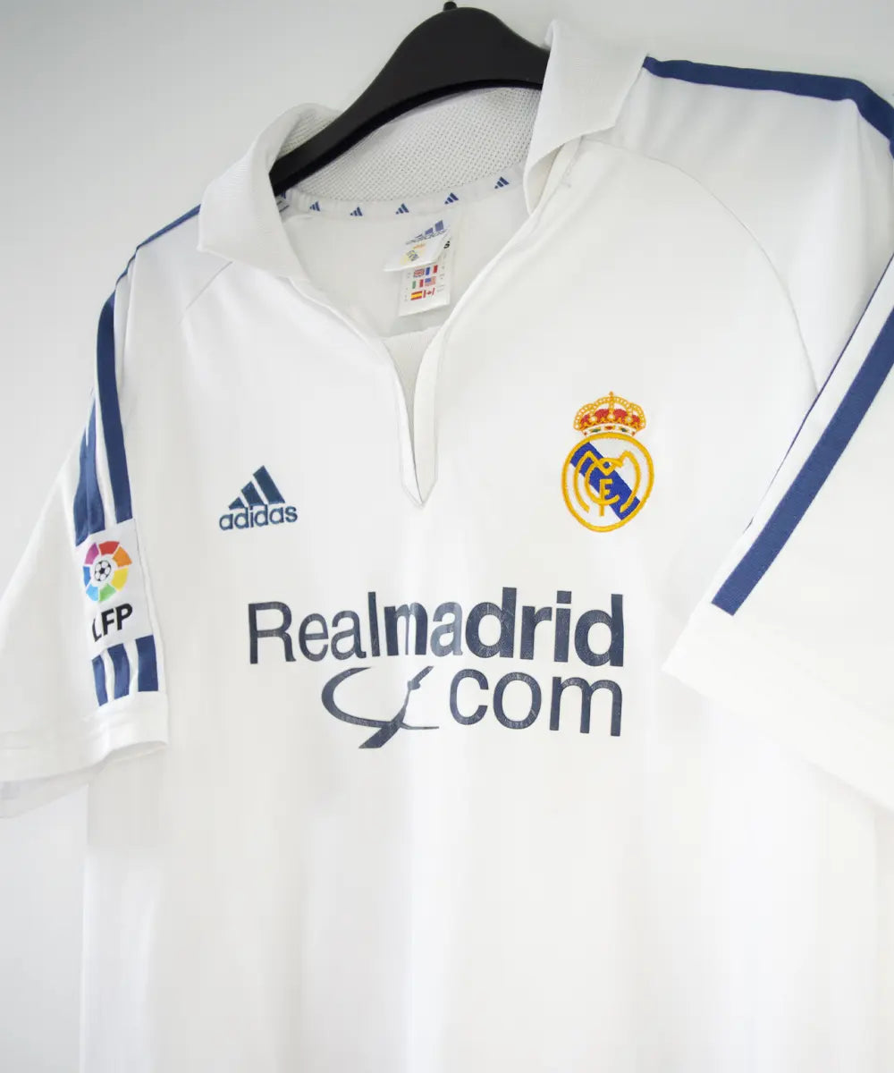 Maillot de foot vintage blanc et bleu du real madrid de la saison 2001-2002. On peut retrouver l'équipementier adidas et le sponsor real madrid.com. Le maillot est flqqué du numéro 5 Zinedine Zidane. Il s'agit d'un maillot authentique.