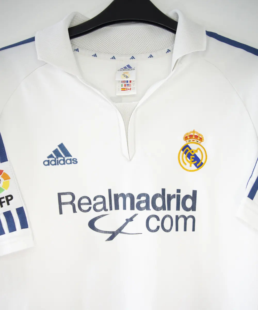 Maillot de foot vintage blanc et bleu du real madrid de la saison 2001-2002. On peut retrouver l'équipementier adidas et le sponsor real madrid.com. Le maillot est flqqué du numéro 5 Zinedine Zidane. Il s'agit d'un maillot authentique.