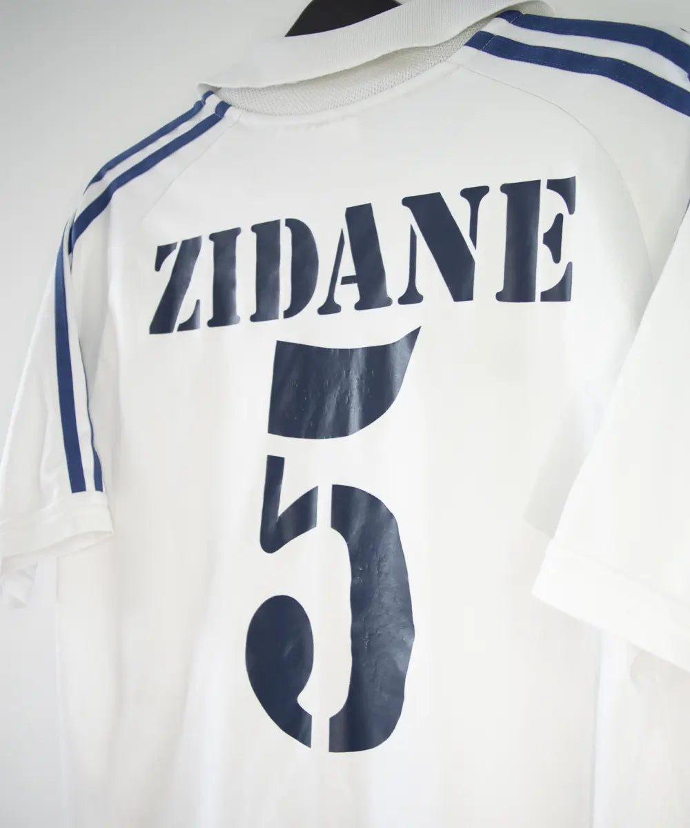 Maillot de foot vintage blanc et bleu du real madrid de la saison 2001-2002. On peut retrouver l'équipementier adidas et le sponsor real madrid.com. Le maillot est flqqué du numéro 5 Zinedine Zidane. Il s'agit d'un maillot authentique.