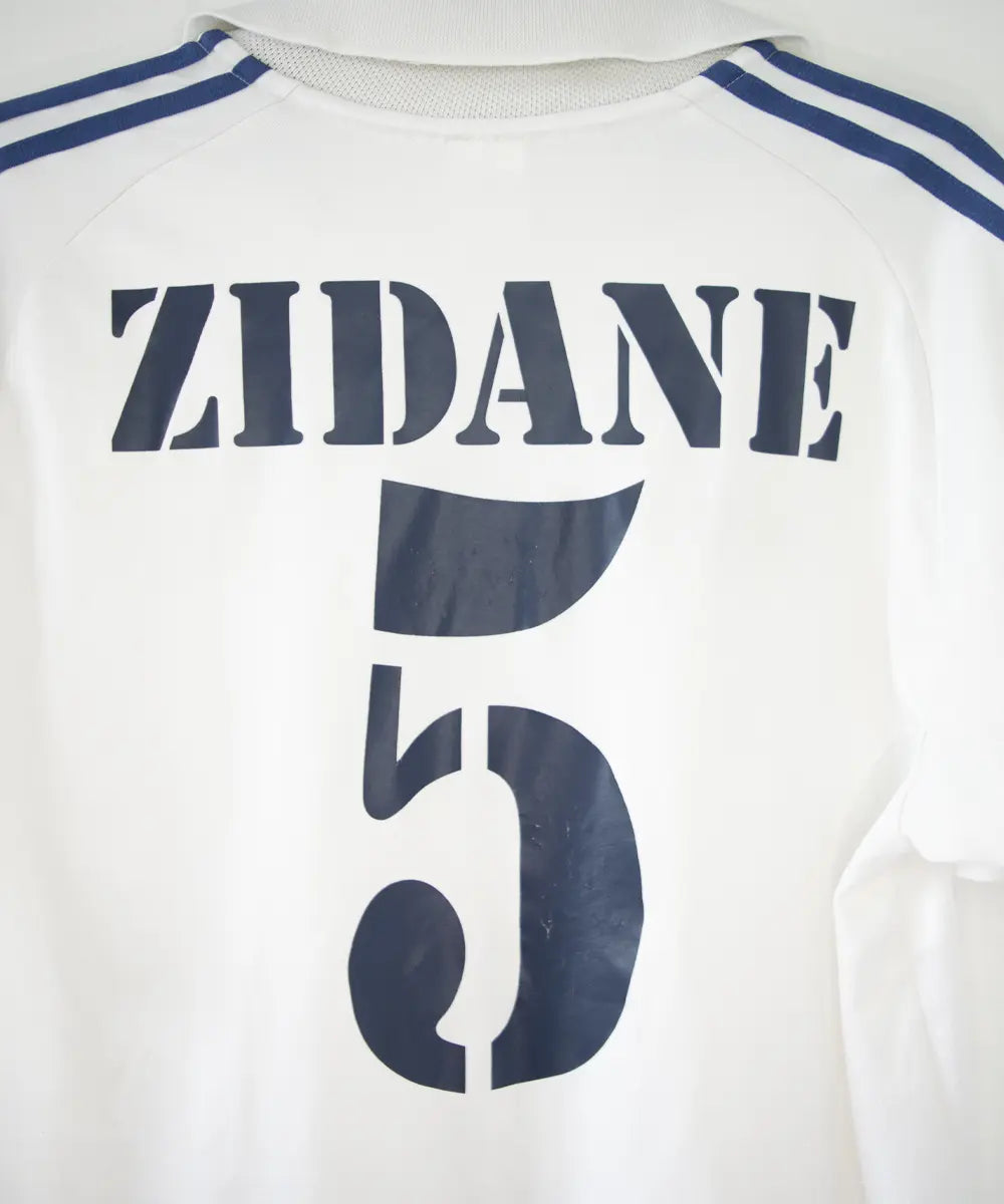 Maillot de foot vintage blanc et bleu du real madrid de la saison 2001-2002. On peut retrouver l'équipementier adidas et le sponsor real madrid.com. Le maillot est flqqué du numéro 5 Zinedine Zidane. Il s'agit d'un maillot authentique.