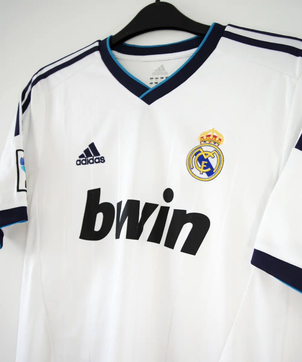 Maillot blanc et bleu du real madrid de la saison 2012-2013. On peut retrouver sur le maillot le sponsor bwin et l'équipementier adidas. Le maillot porte aussi le patch des 110 ans du real et le patch de la LFP. Sur cette photo on peut voir le devant du maillot de coté