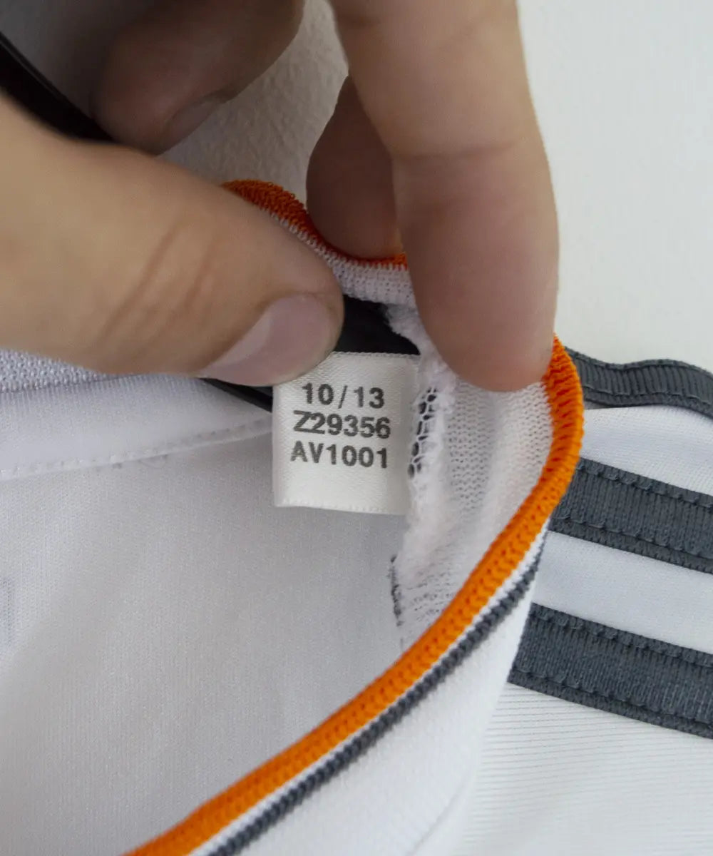 Maillot domicile blanc du réal madrid de la saison 2013-2014. On peut retrouver l'équipementier adidas et le sponsor fly emirates. Le maillot est floqué du numéro 7 Ronaldo. Sur cette photo on peut voir l'étiquette du maillot comportant les numéros Z29356