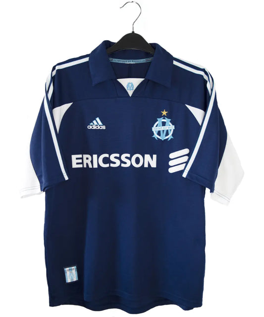 Maillot vintage extérieur bleu et blanc de l'om de la saison 1999-2000. On peut retrouver l'équipementier adidas et le sponsor ericsson