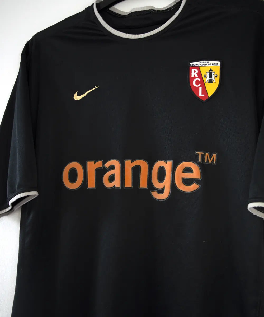 Maillot extérieur du RC Lens de la saison 2002-2003 de couleur noir et blanc. On peut retrouver l'équipementier nike et le sponsor orange.