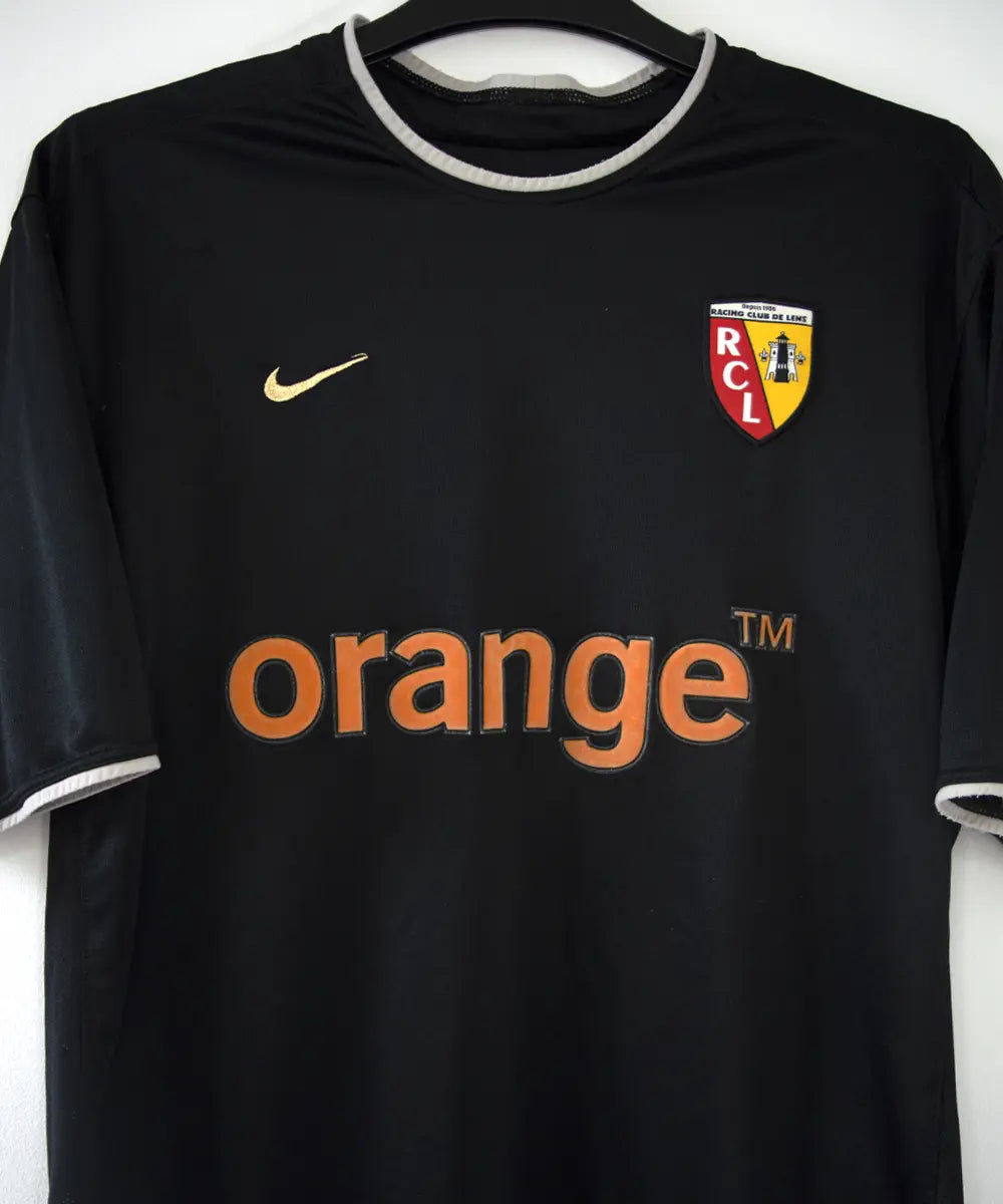 Maillot extérieur du RC Lens de la saison 2002-2003 de couleur noir et blanc. On peut retrouver l'équipementier nike et le sponsor orange.
