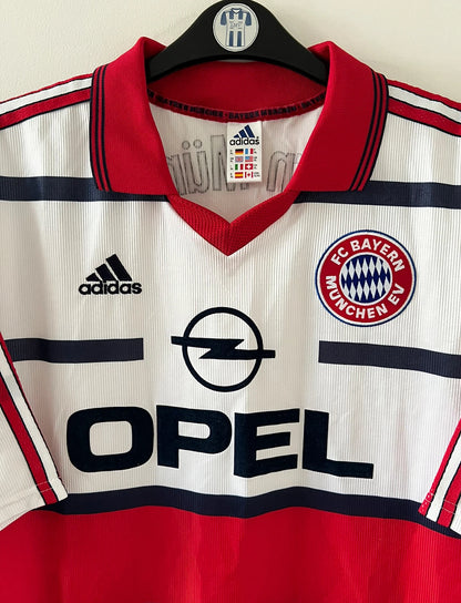 Maillot de foot vintage extérieur blanc et rouge du Bayern Munich 1998/2000. On peut retrouver l'équipementier Adidas et le sponsor Opel. Il s'agit d'un maillot authentique d'époque.