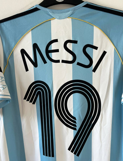 Maillot de foot vintage domicile de l'Argentine 2006. On peut retrouver l'équipementier Adidas. Le maillot est floqué du numéro 19 Messi. Il s'agit d'un maillot authentique d'époque.