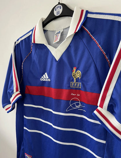 Maillot de foot vintage bleu, blanc et rouge de l'équipe de france 1998. On peut retrouver l'équipementier adidas. Il s'agit d'un maillot authentique édité avant la finale de la coupe du monde car celui-ci ne possède pas l'étoile. Le maillot possède la mention pour toi avec la signature de zidane