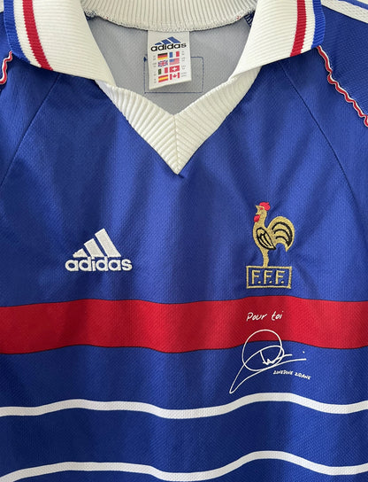 Maillot de foot vintage bleu, blanc et rouge de l'équipe de france 1998. On peut retrouver l'équipementier adidas. Il s'agit d'un maillot authentique édité avant la finale de la coupe du monde car celui-ci ne possède pas l'étoile. Le maillot possède la mention pour toi avec la signature de zidane