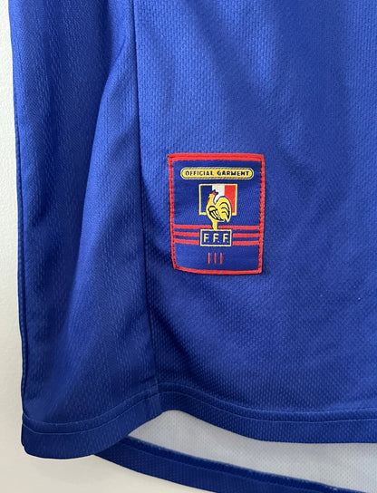 Maillot de foot vintage bleu, blanc et rouge de l'équipe de france 1998. On peut retrouver l'équipementier adidas. Il s'agit d'un maillot authentique édité avant la finale de la coupe du monde car celui-ci ne possède pas l'étoile. Le maillot possède la mention pour toi avec la signature de zidane