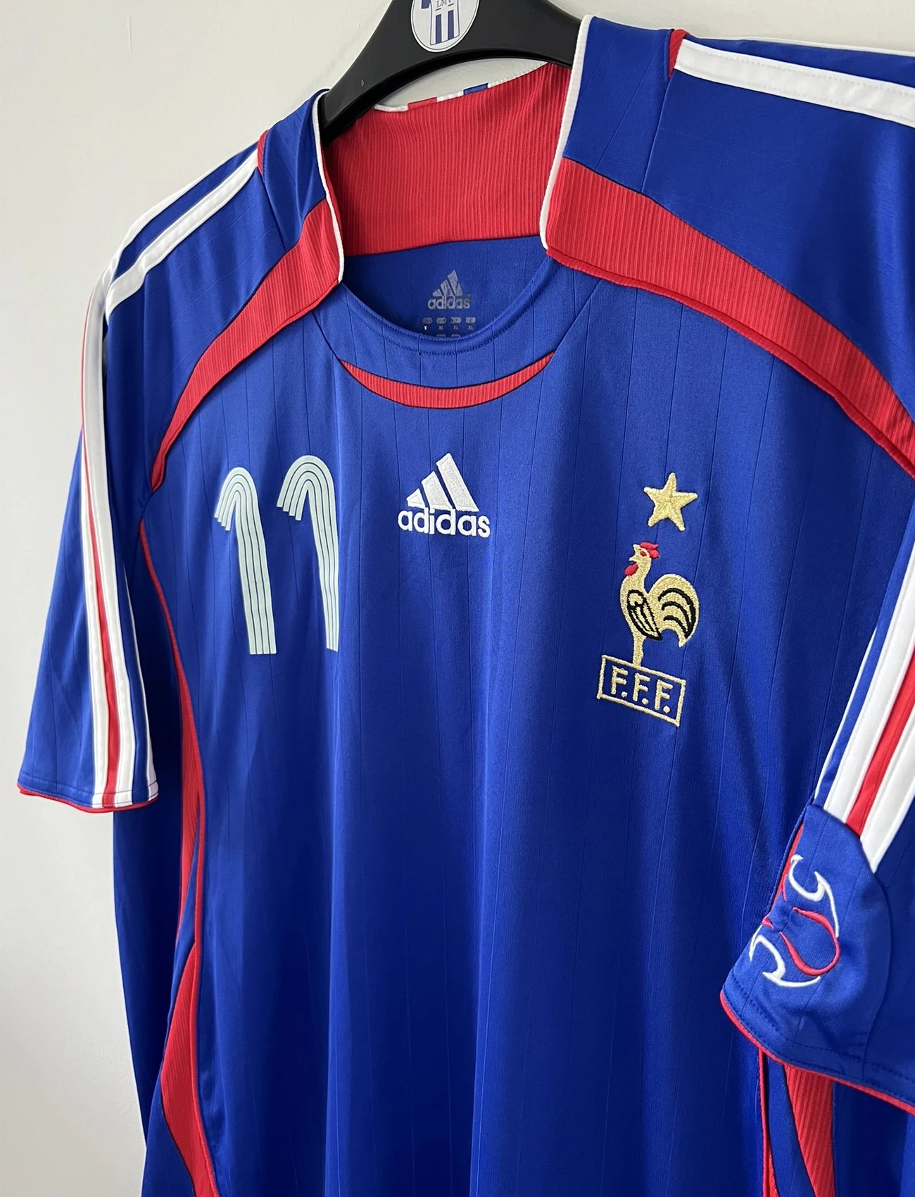 Maillot de foot vintage domicile bleu, blanc et rouge de l'équipe de france 2006. On peut retrouver l'équipementier adidas. Le maillot est floqué du numéro 11 Sylvain Wiltord. Il s'agit d'un maillot authentique d'époque comportant l'étiquette 740126