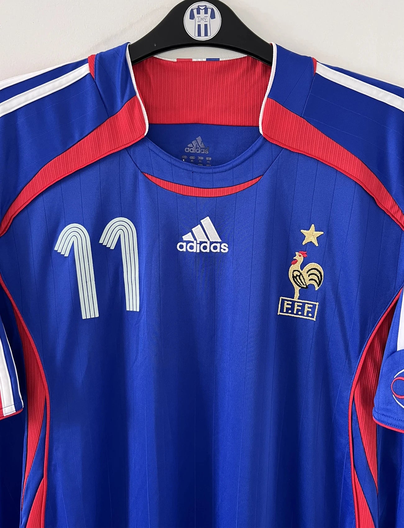 Maillot de foot vintage domicile bleu, blanc et rouge de l'équipe de france 2006. On peut retrouver l'équipementier adidas. Le maillot est floqué du numéro 11 Sylvain Wiltord. Il s'agit d'un maillot authentique d'époque comportant l'étiquette 740126