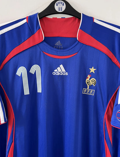 Maillot de foot vintage domicile bleu, blanc et rouge de l'équipe de france 2006. On peut retrouver l'équipementier adidas. Le maillot est floqué du numéro 11 Sylvain Wiltord. Il s'agit d'un maillot authentique d'époque comportant l'étiquette 740126