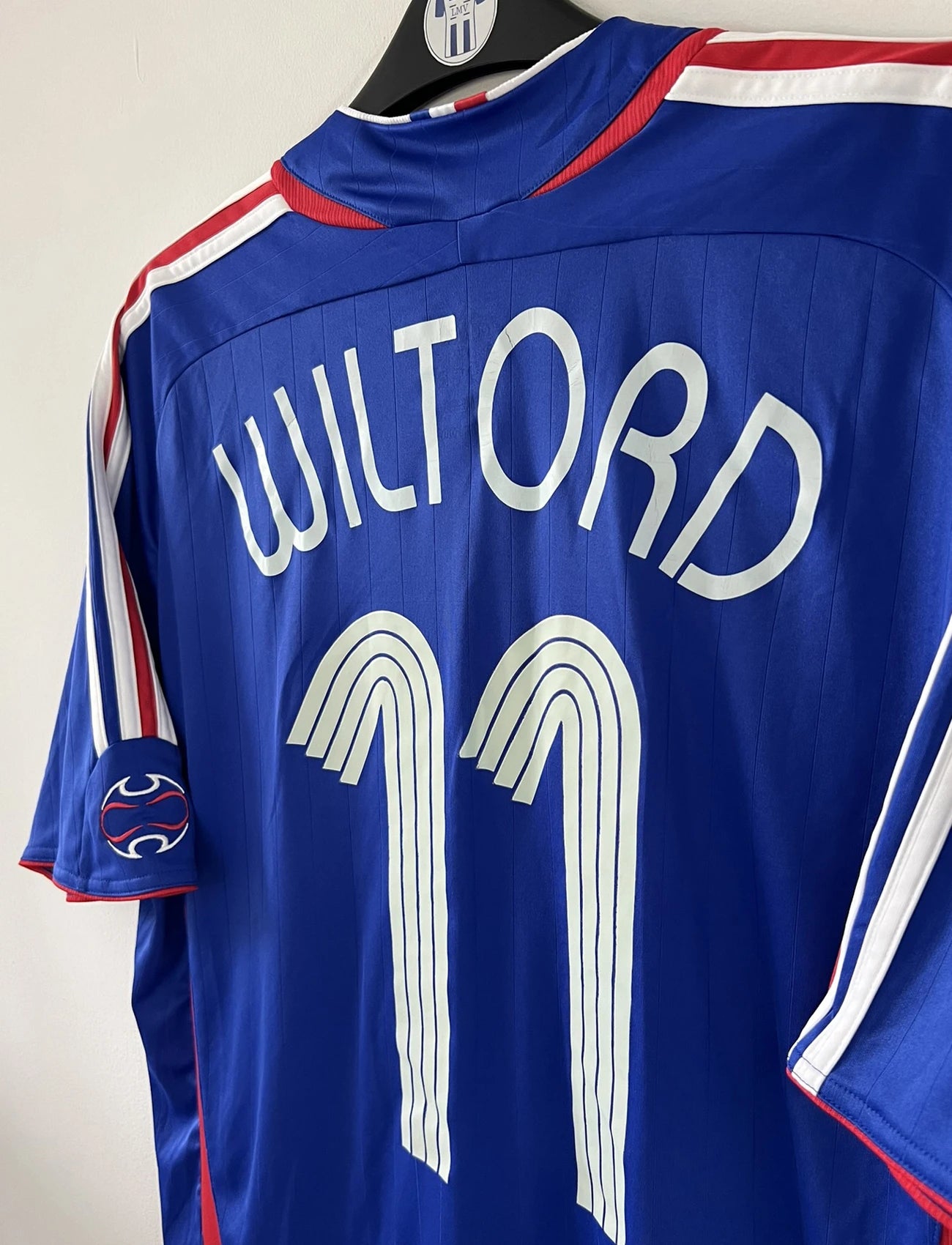 Maillot de foot vintage domicile bleu, blanc et rouge de l'équipe de france 2006. On peut retrouver l'équipementier adidas. Le maillot est floqué du numéro 11 Sylvain Wiltord. Il s'agit d'un maillot authentique d'époque comportant l'étiquette 740126