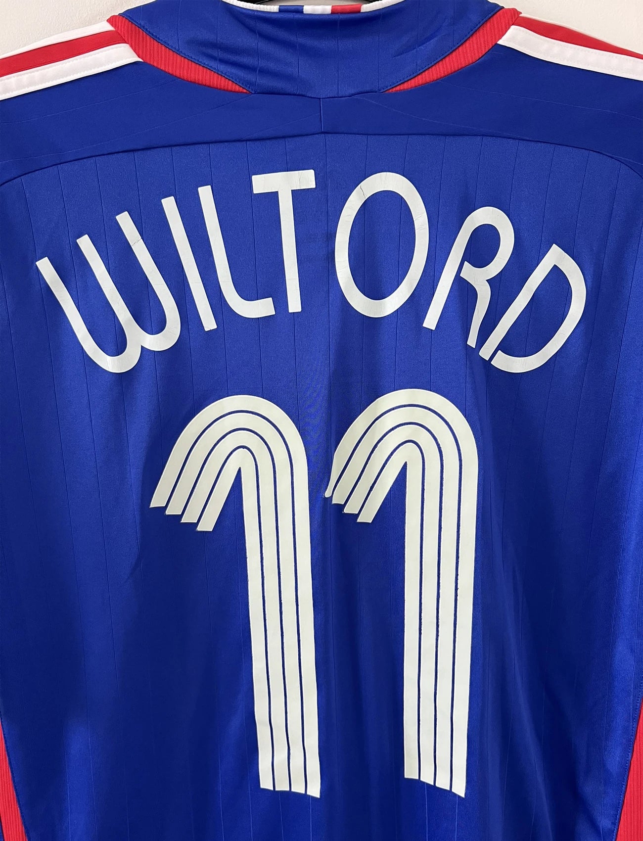 Maillot de foot vintage domicile bleu, blanc et rouge de l'équipe de france 2006. On peut retrouver l'équipementier adidas. Le maillot est floqué du numéro 11 Sylvain Wiltord. Il s'agit d'un maillot authentique d'époque comportant l'étiquette 740126
