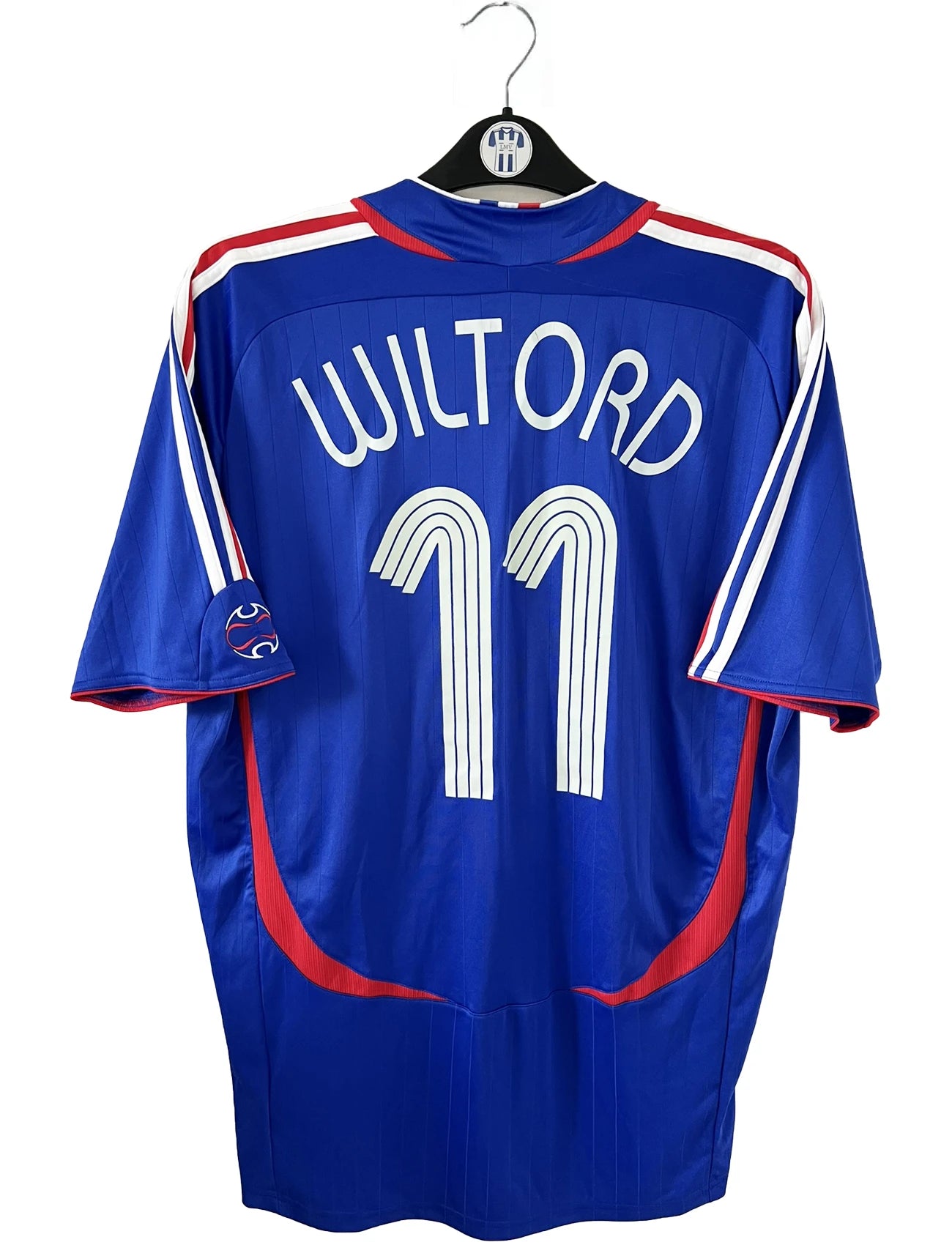 Maillot de foot vintage domicile bleu, blanc et rouge de l'équipe de france 2006. On peut retrouver l'équipementier adidas. Le maillot est floqué du numéro 11 Sylvain Wiltord. Il s'agit d'un maillot authentique d'époque comportant l'étiquette 740126