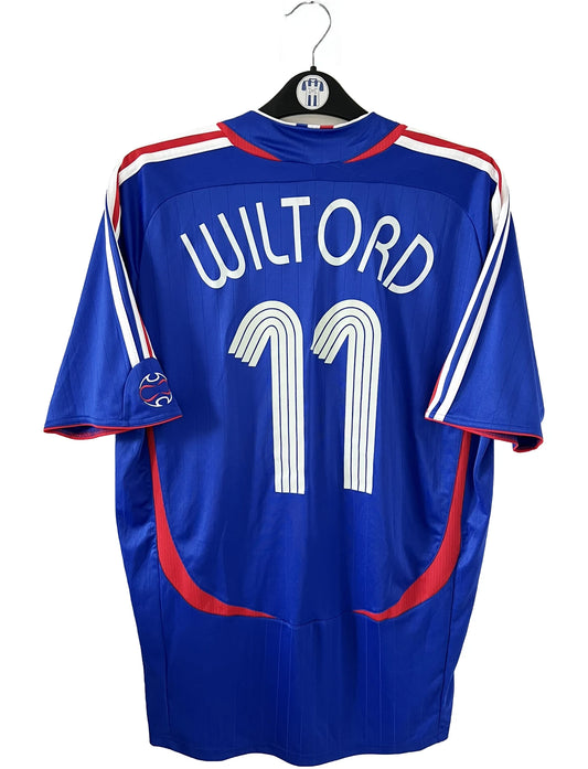 Maillot de foot vintage domicile bleu, blanc et rouge de l'équipe de france 2006. On peut retrouver l'équipementier adidas. Le maillot est floqué du numéro 11 Sylvain Wiltord. Il s'agit d'un maillot authentique d'époque comportant l'étiquette 740126