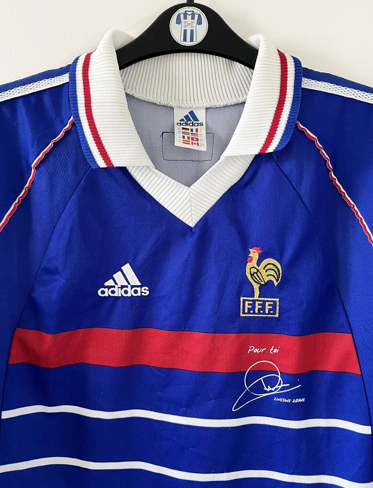 Maillot de foot vintage bleu, blanc et rouge de l'équipe de france 1998. On peut retrouver l'équipementier adidas. Il s'agit d'un maillot authentique édité avant la finale de la coupe du monde car celui-ci ne possède pas l'étoile. Le maillot possède la mention pour toi avec la signature de zidane