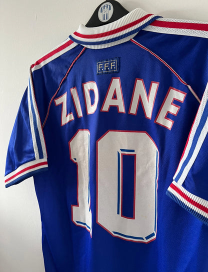 Maillot de foot vintage domicile bleu blanc et rouge de l'équipe de france 1998. On peut retrouver l'équipementier Adidas. Le maillot est floqué du numéro 10 Zinedine Zidane. Il s'agit d'un maillot authentique d'époque.
