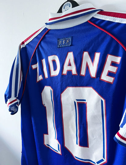 Maillot de foot vintage domicile bleu de l'équipe de France 1998. On peut retrouver l'équipementier Adidas, la signature et le flocage de Zinedine Zidane. Il s'agit d'une magnifique pièce de collection.
