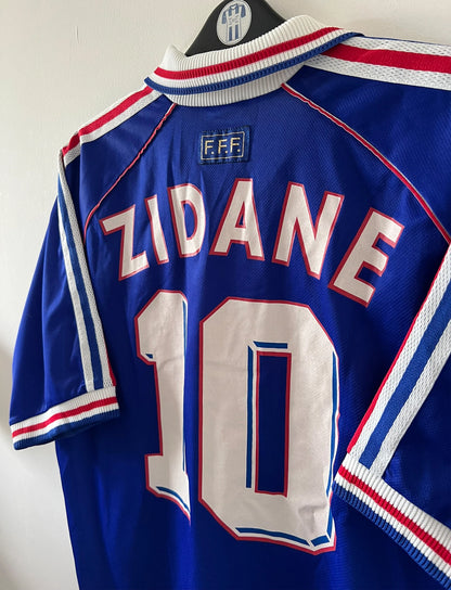 Maillot de foot vintage domicile bleu de l'équipe de france 1998. On peut retrouver l'équipementier Adidas. Le maillot est floqué du numéro 10 Zinedine Zidane. Il s'agit d'un maillot authentique d'époque.