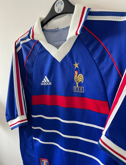 Maillot de foot vintage domicile bleu de l'équipe de france 1998. On peut retrouver l'équipementier Adidas. Le maillot est floqué du numéro 10 Zinedine Zidane. Il s'agit d'un maillot authentique d'époque.