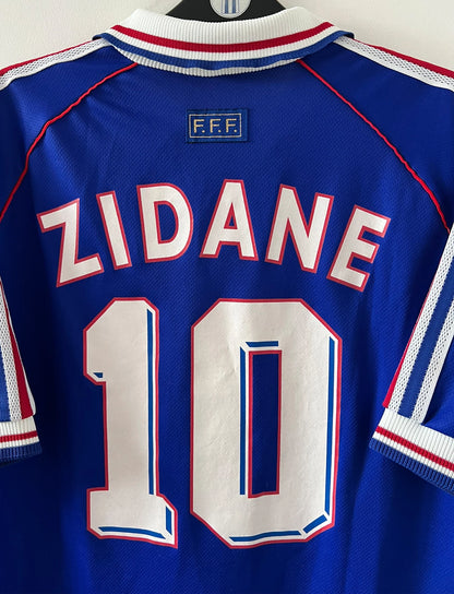 Maillot de foot vintage domicile bleu de l'équipe de france 1998. On peut retrouver l'équipementier Adidas. Le maillot est floqué du numéro 10 Zinedine Zidane. Il s'agit d'un maillot authentique d'époque.