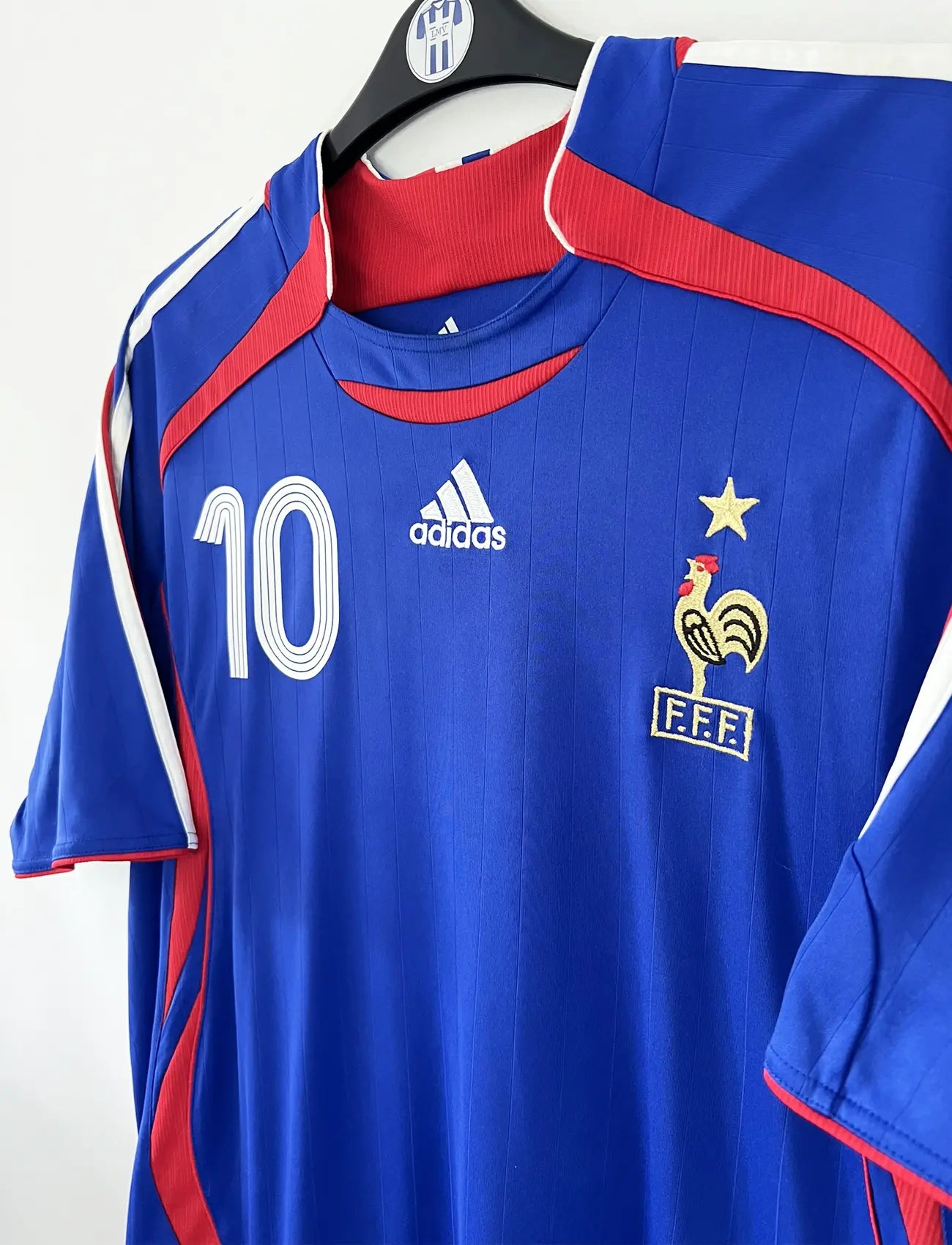 Maillot de foot vintage domicile bleu, blanc et rouge de l'équipe de france 2006. On peut retrouver l'équipementier adidas. Le maillot est floqué du numéro 10 Zinedine Zidane. Il s'agit d'un maillot authentique d'époque comportant l'étiquette 740126. Le maillot est un taille XL