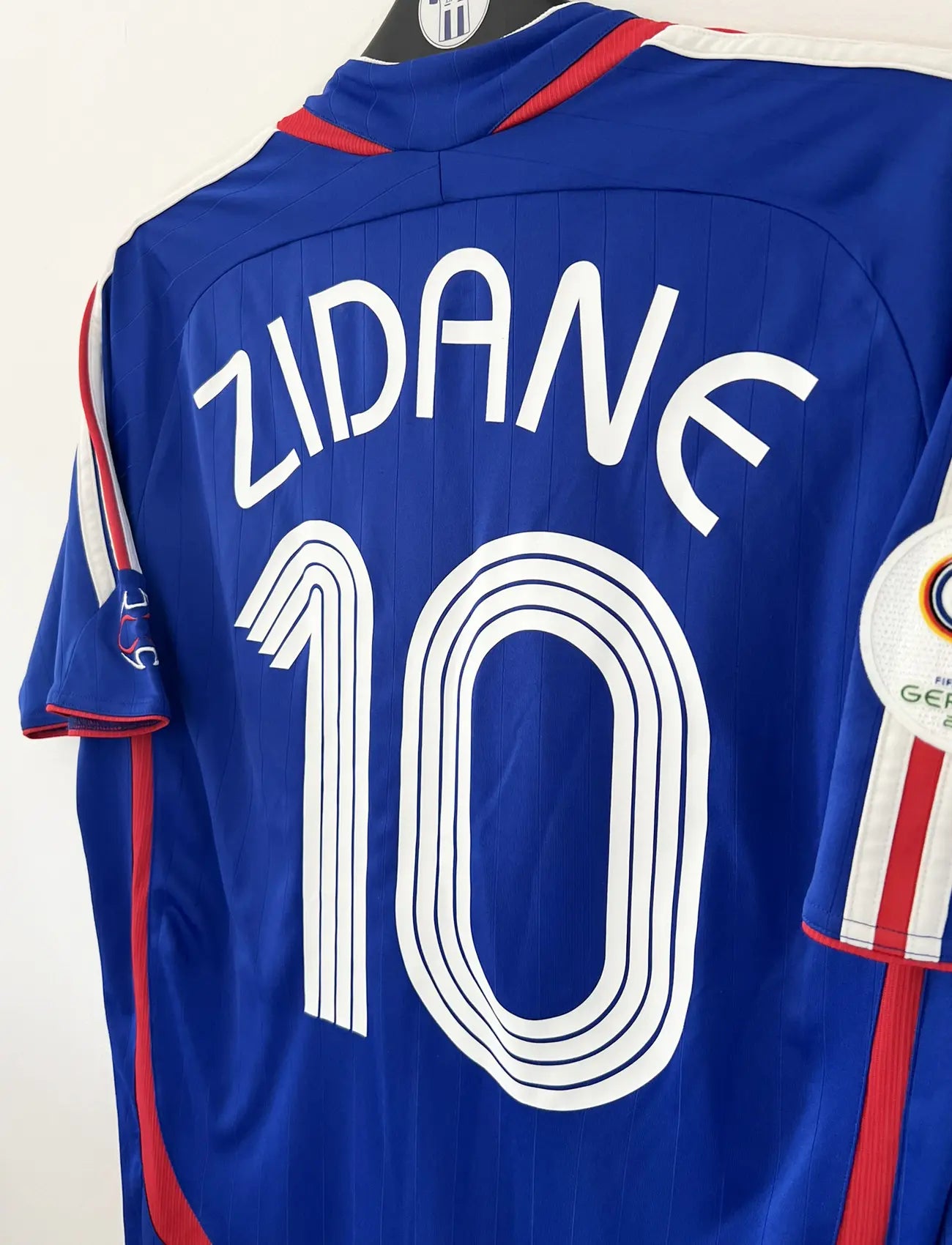 Maillot de foot vintage domicile bleu, blanc et rouge de l'équipe de france 2006. On peut retrouver l'équipementier adidas. Le maillot est floqué du numéro 10 Zinedine Zidane. Il s'agit d'un maillot authentique d'époque comportant l'étiquette 740126