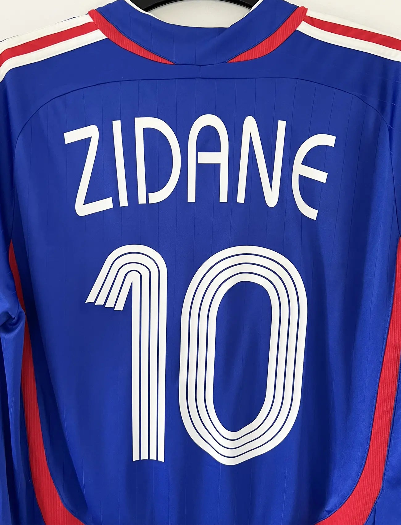 Maillot de foot vintage domicile bleu, blanc et rouge de l'équipe de france 2006. On peut retrouver l'équipementier adidas. Le maillot est floqué du numéro 10 Zinedine Zidane. Il s'agit d'un maillot authentique d'époque comportant l'étiquette 740126. Le maillot est un taille XL