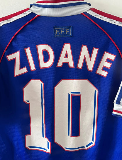 Maillot foot vintage domicile equipe de france 1998 bleu blanc et rouge. On peut retrouver l'équipementier adidas. Il s'agit d'un maillot authentique d'époque. Le maillot est floqué du numéro 10 Zinedine Zidane et comporte également sa signature avec la mention "pour toi Zinedine Zidane"