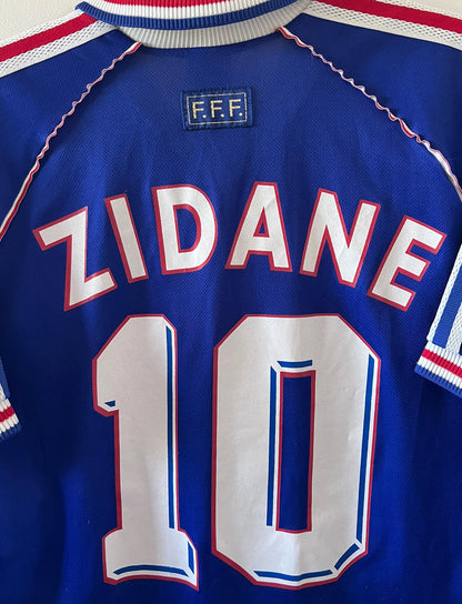 Maillot de foot vintage domicile bleu de l'équipe de france 1998. On peut retrouver l'équipementier Adidas. Le maillot est floqué du numéro 10 Zinedine Zidane. Il s'agit d'un maillot authentique d'époque.