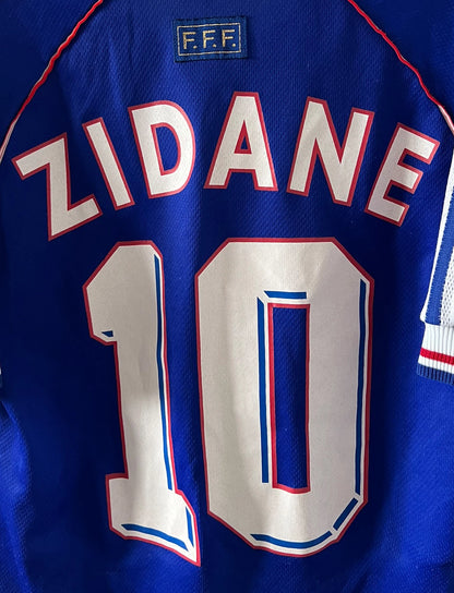Maillot de foot vintage domicile bleu de l'équipe de france 1998. On peut retrouver l'équipementier Adidas. Le maillot est floqué du numéro 10 Zinedine Zidane. Il s'agit d'un maillot authentique d'époque.