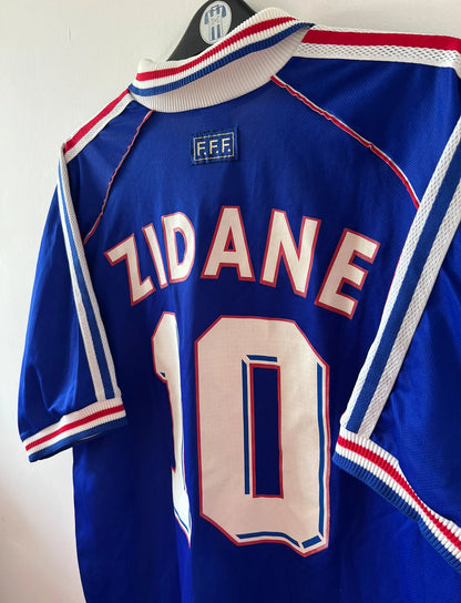 Maillot de foot vintage domicile bleu de l'équipe de France 1998. On peut retrouver l'équipementier Adidas. Le maillot est floqué du numéro 10 Zinedine Zidane. Il s'agit d'un maillot authentique d'époque.