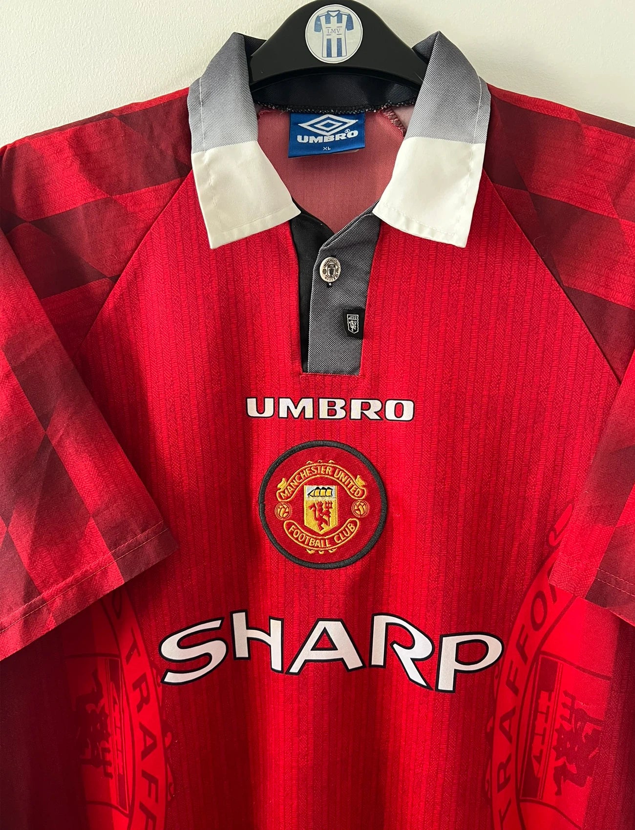 Maillot de foot vintage domicile rouge de Manchester United 1996/1998. On peut retrouver l'équipementier Umbro et le sponsor Sharp. Le maillot est floqué du numéro 7 Eric Cantona. Il s'agit d'un maillot authentique d'époque.