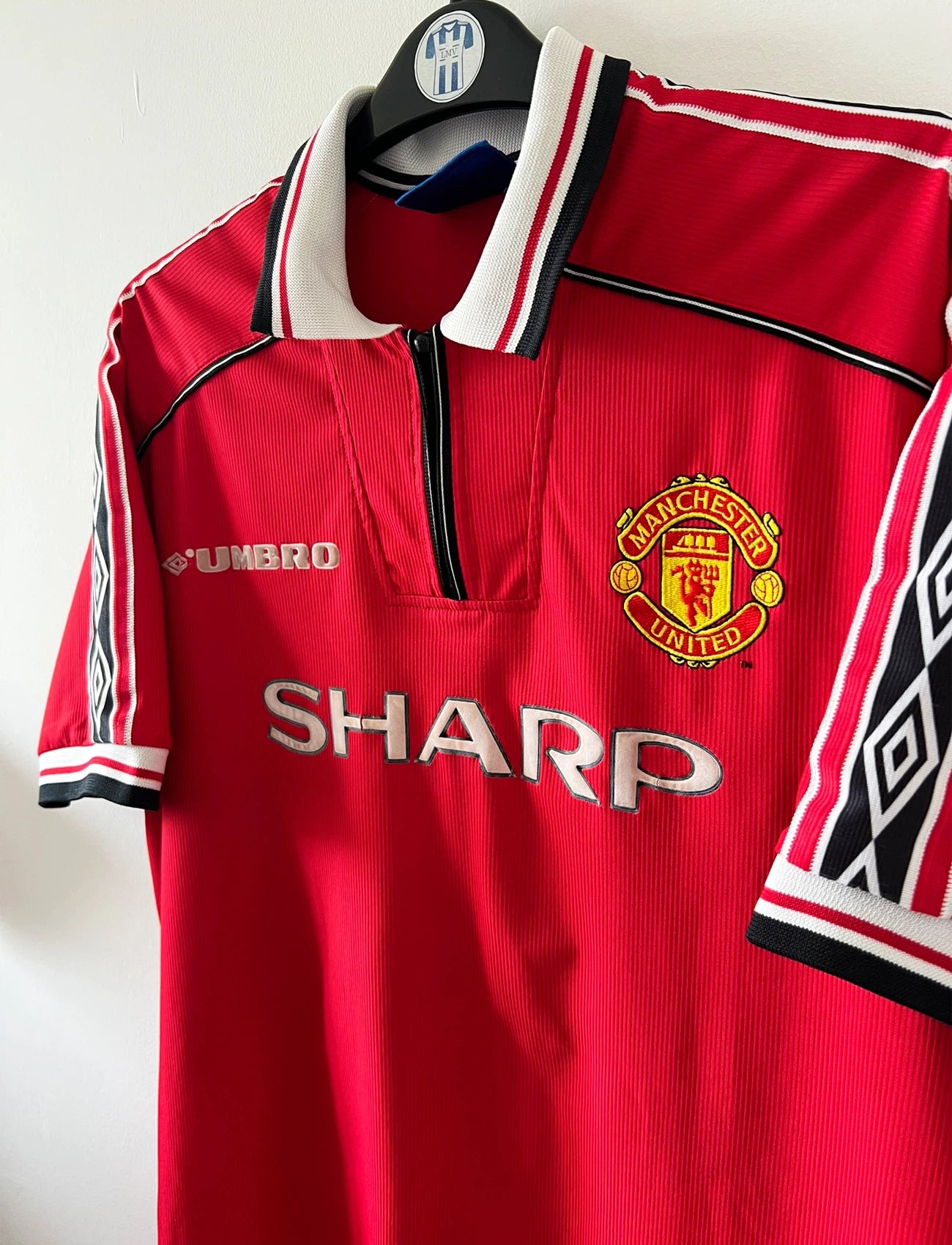 Maillot foot vintage domicile rouge de Manchester United 1998. On peut retrouver l'équipementier Umbro et le sponsor Sharp. Il s'agit d'un maillot authentique d'époque.