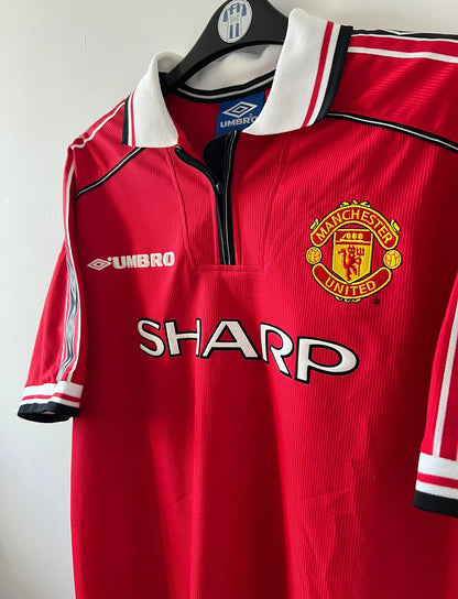 Maillot de foot vintage domicile rouge de Manchester United saison 1998/2000. On peut retrouver l'équipementier Umbro et le sponsor Sharp. Il s'agit d'un maillot authentique d'époque.
