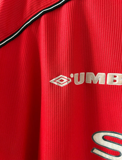 Maillot foot vintage domicile rouge de Manchester United 1998. On peut retrouver l'équipementier Umbro et le sponsor Sharp. Il s'agit d'un maillot authentique d'époque.