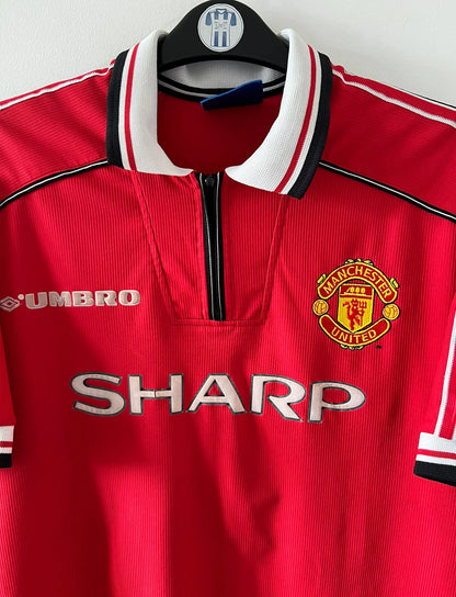 Maillot foot vintage domicile rouge de Manchester United 1998. On peut retrouver l'équipementier Umbro et le sponsor Sharp. Il s'agit d'un maillot authentique d'époque.