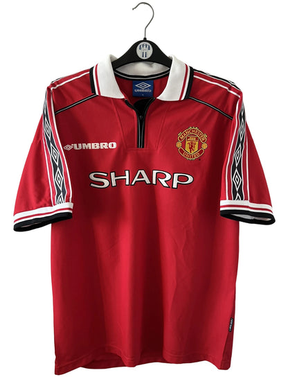 Maillot de foot vintage domicile rouge de Manchester United saison 1998/2000. On peut retrouver l'équipementier Umbro et le sponsor Sharp. Il s'agit d'un maillot authentique d'époque.