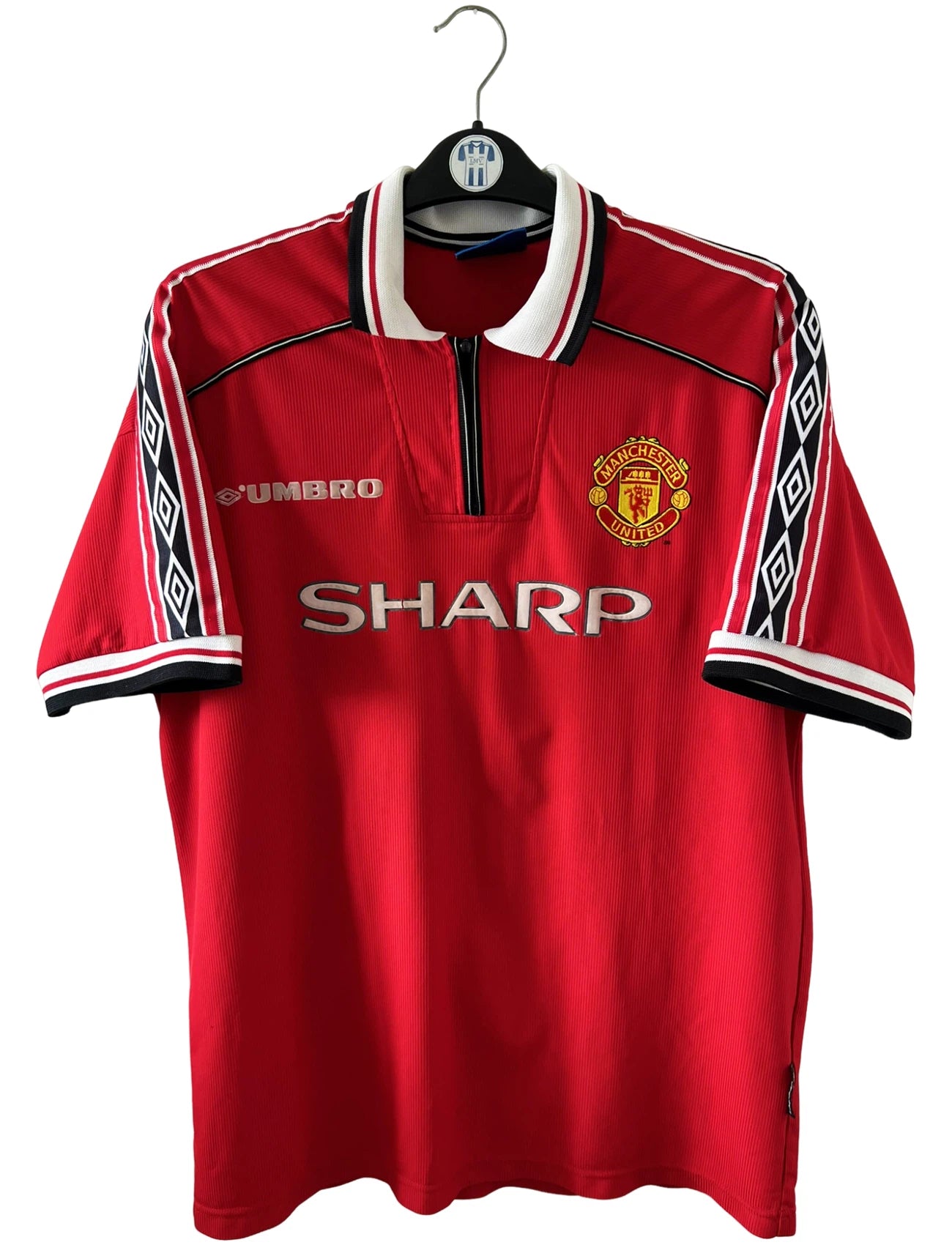 Maillot foot vintage domicile rouge de Manchester United 1998. On peut retrouver l'équipementier Umbro et le sponsor Sharp. Il s'agit d'un maillot authentique d'époque.