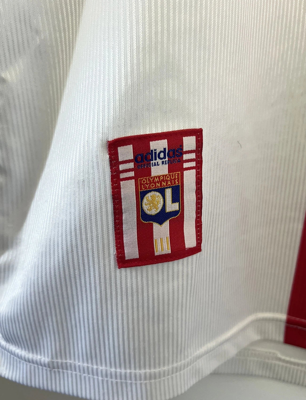 Maillot de foot vintage domicile blanc, bleu et rouge de l'Olympique Lyonnais de la saison 1998/2000. On peut retrouver l'équipementier Adidas et le sponsor Infogrames. Il s'agit d'un maillot authentique d'époque.