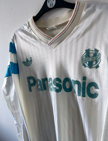 Maillot foot vintage domicile blanc de l'OM 1989/1991. On peut retrouver l'équipementier Adidas et le sponsor Panasonic. Il s'agit d'un maillot authentique d'époque.
