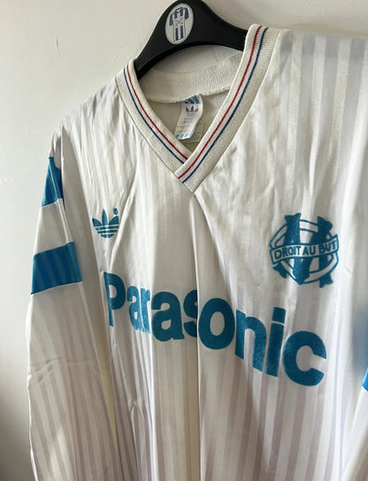 Maillot de foot vintage domicile blanc et bleu de l'om 1989/1991. On peut retrouver l'équipementier Adidas et le sponsor Panasonic. Il s'agit d'un maillot authentique d'époque.