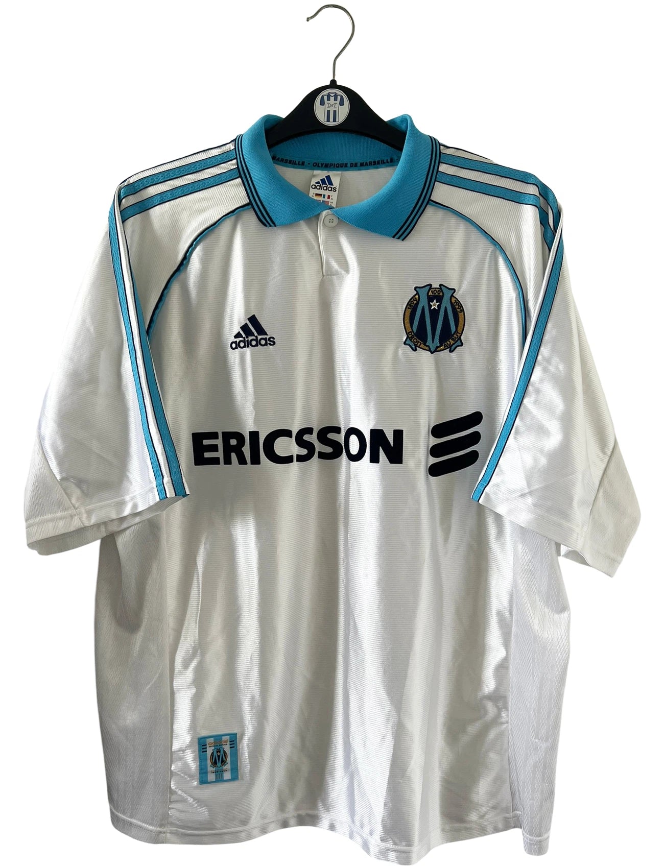 Maillot de foot vintage domicile blanc et bleu de l'OM de la saison 1998-1999. On peut retrouver l'équipementier Adidas et le sponsor Ericsson. Il s'agit d'un maillot authentique d'époque.