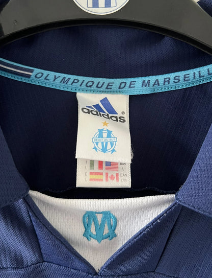 Maillot de foot vintage OM 1999/2000 extérieur bleu. On peut retrouver l'équipementier Adidas et le sponsor Ericsson. Il s'agit d'un maillot authentique d'époque.