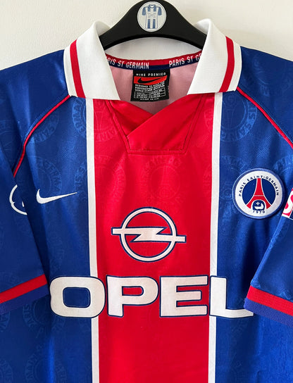 Maillot de foot vintage domicile rouge et bleu du PSG de la saison 1996/1997. On peut retrouver la fameuse bande hechter avec l'équipementier Nike et le sponsor Opel. Il s'agit d'un maillot authentique d'époque.