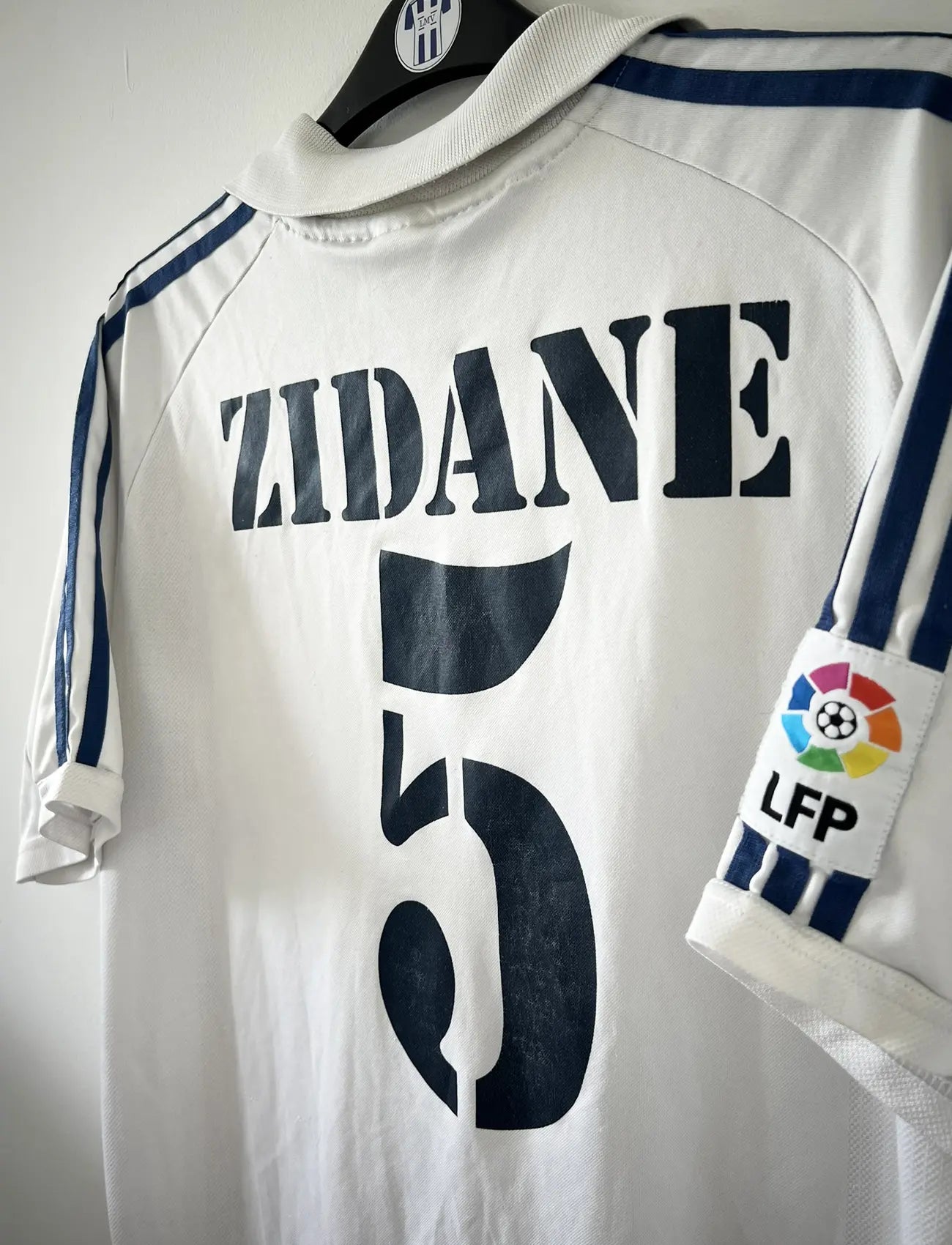 Maillot de foot vintage domicile du real madrid de la saison 2001-2002. Le maillot est de couleur blanc et noir. On peut retrouver l'équipementier adidas et le sponsor Real Madrid.com. Le maillot est floqué du numéro 5 Zinedine Zidane. Il s'agit d'un maillot authentique comportant les numéros 695856