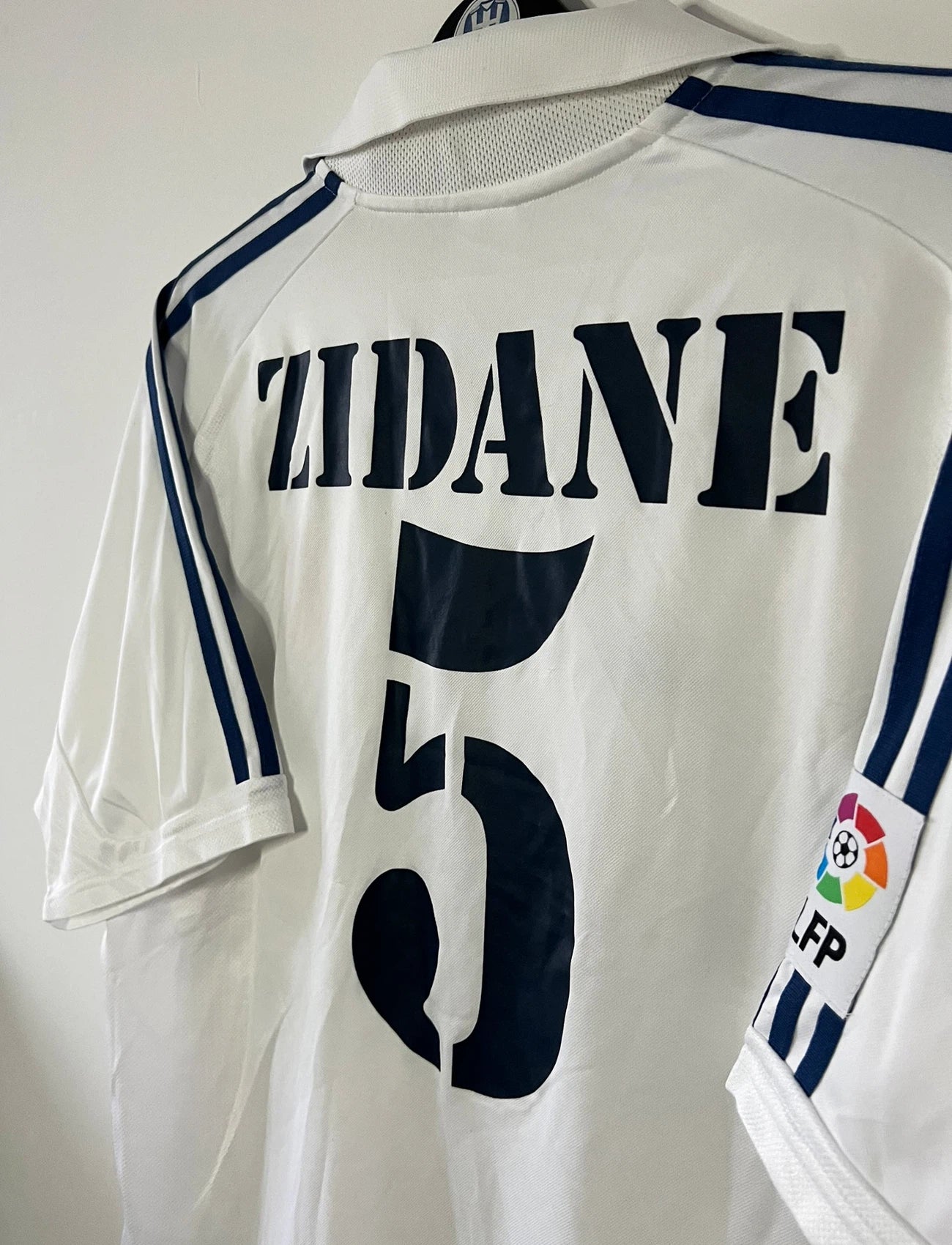 Maillot de foot vintage domicile blanc du real madrid de la saison 2001/2002. On peut retrouver l'équipementier Adidas et le sponsor Real madrid.com. Le maillot est floqué du numéro 5 Zinedine Zidane. Il s'agit d'un maillot authentique d'époque.